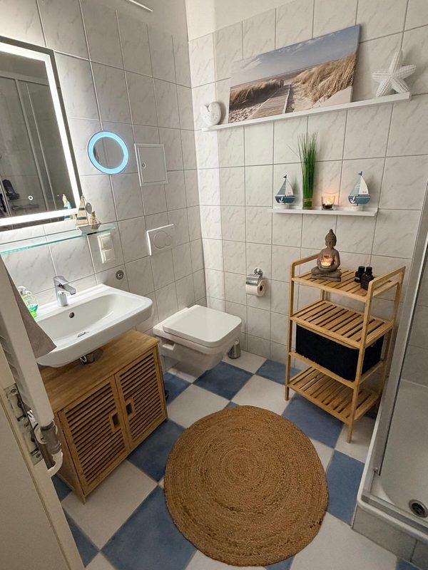 Badezimmer mit Waschtisch, beleuchtetem Spiegel, WC und Holzregal, Boden blau-weiß gefliest