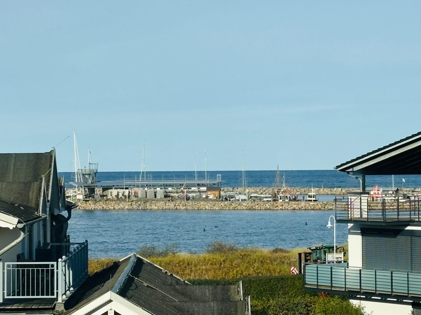 Blick zwischen zwei Häusern auf den Hafen von Glowe mit Segelmasten und Mole, dahinter die Ostsee