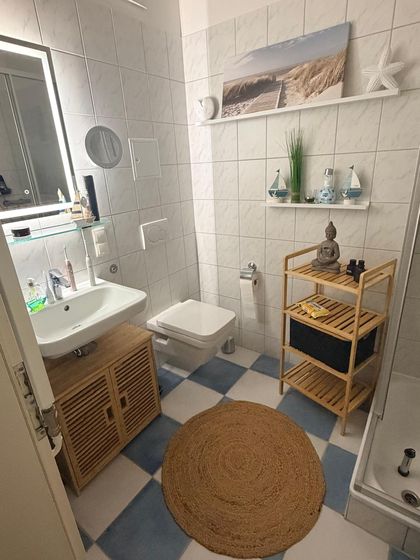 Badezimmer mit Waschtisch, beleuchtetem Spiegel, WC und Holzregal, blau-weiß gefliester Boden